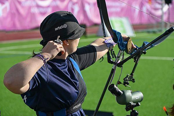Eine Schützin zielt mit einem Bogen auf ein Ziel, während sie auf einer grünen Sportfläche steht.