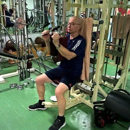 Mann trainiert mit einem Fitnessgerät im Gym, während im Hintergrund weitere Sportgeräte und Personen sichtbar sind.