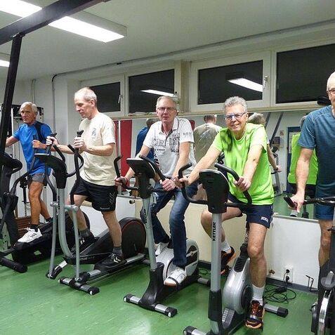 Fünf Männer trainieren auf Crosstrainern in einem Fitnessraum mit grüner Bodenbelag und großen Fenstern.