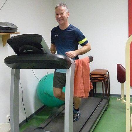 Mann joggt lächelnd auf einem Laufband in einem Fitnessraum, ein Handtuch über der Schulter. Grüne Bodenfläche sichtbar.