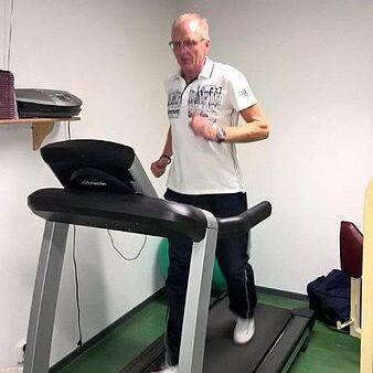 Älterer Mann joggt auf einem Laufband in einem hellen Raum mit grünem Boden und Geräten im Hintergrund.