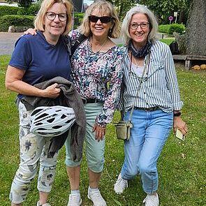 Drei Frauen lächeln und stehen arm in arm auf einer Wiese, umgeben von Bäumen und Verkehrsschildern.