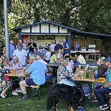 Gruppe von Menschen sitzt an Tischen vor einem Imbissstand unter Bäumen, darunter ein Hund. Viele sind in angeregter Unterhaltung.