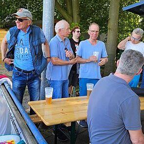 Fünf Personen unterhalten sich an einer Tischtennisanlage, einige halten Getränke in der Hand. Bäume im Hintergrund.