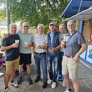 Gruppe von sechs Männern, die lächelnd mit Getränken in der Hand vor einem Kiosk stehen, umgeben von Bäumen.