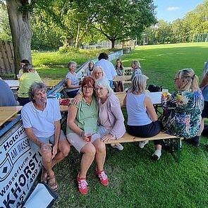 Drei Frauen sitzen an einer Holzbank im Freien, umgeben von weiteren Leuten und Bäumen auf einer Wiese.
