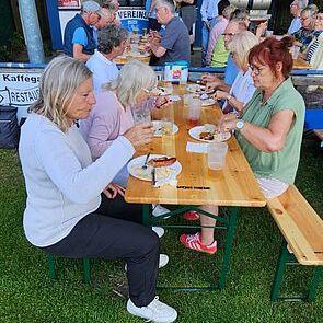 Gruppe von Menschen sitzt an Tischen im Freien, genießt Essen und Getränke vor einem Vereinsgebäude.