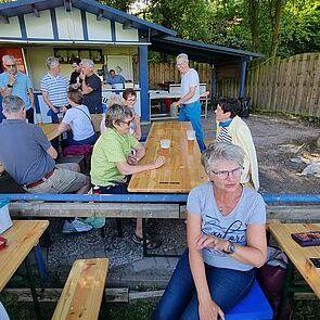 Eine Gruppe von Menschen sitzt und steht um Holztische auf einer Freifläche vor einem Gebäude.