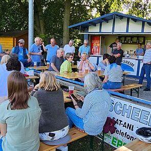 Eine Gruppe von Menschen in lässiger Kleidung versammelt sich um Bänke vor einer kleinen Hütte im Freien.