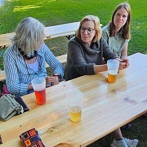 Drei Frauen sitzen an einem Holztisch im Freien, mit Getränken in plastikbechern und einer Wiese im Hintergrund.
