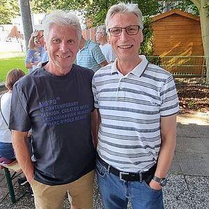 Zwei Männer stehen nebeneinander in einem Garten, umgeben von weiteren Personen im Hintergrund.