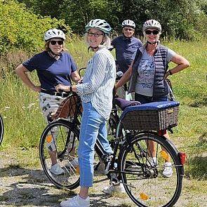 Vier Frauen mit Fahrradhelmen posieren freundlich im Freien, während eine Frau auf ihrem Fahrrad steht.