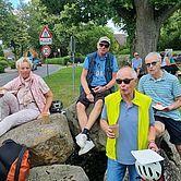 Vier Männer sitzen entspannt auf Steinen, genießen Snacks und Getränke, umgeben von Grünanlagen und Straßenverkehrsschildern.