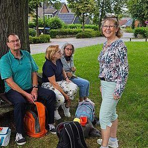 Vier Personen sitzen unter einem Baum auf einer Wiese, lächelnd, während eine Person aufsteht und in die Kamera schaut.