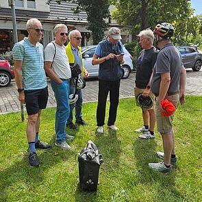 Gruppe von sechs Männern in Radbekleidung steht im Freien, diskutiert und trägt Helme auf einer Wiese.