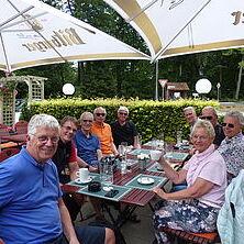 Gruppe von acht Personen sitzt an einem Tisch unter Sonnenschirmen in einem Gartenrestaurant, Tische mit Getränken und Speisen.