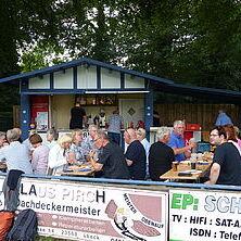 Gruppe von Personen sitzt an Tischen vor einem Imbissstand, während andere drinnen bestellen. Bäume im Hintergrund.