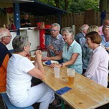 Gruppe von sechs Personen sitzt an einem Holztisch, trinkend und im Gespräch, im Freien bei einem geselligen Treffen.