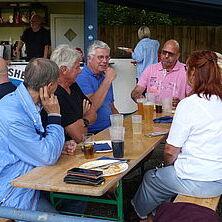 Gruppe von sieben Personen an einem Tisch, Unterhaltungen und Getränke auf Holzbank im Freien.