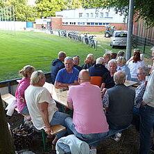 Gruppe von Personen sitzt an einem Tisch im Freien, im Hintergrund eine Wiese und Gebäude.