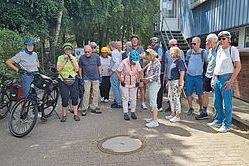 Gruppe von Radfahrern in Sportkleidung und Helmen versammelt vor einem Gebäude, einige mit Fahrrädern, im Freien.
