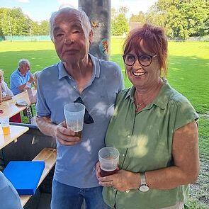 Zwei Personen stehen unter einem Pavillon, halten Kunststoffbecher und posieren vor einem grünen Rasenfeld.