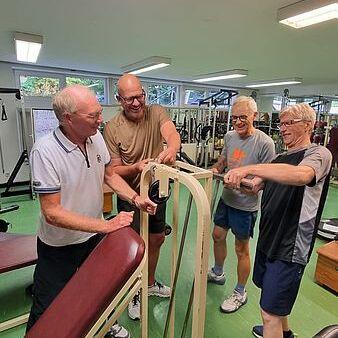Vier ältere Männer in einem Fitnessstudio stehen um ein Trainingsgerät und diskutieren dessen Nutzung.
