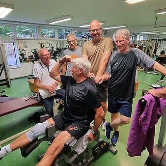 Mehrere Männer im Fitnessstudio beobachten und unterstützen einen älteren Mann beim Training an einem Beintrainer.