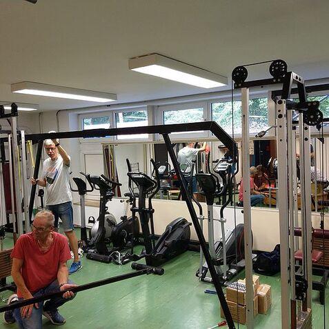 Zwei Männer im Fitnessstudio, einer kniet und hält eine Stange, der andere steht und justiert Geräte.