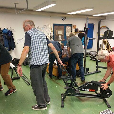 Eine Gruppe von Männern arrangiert Fitnessgeräte in einem Raum mit grünem Boden und Wänden.