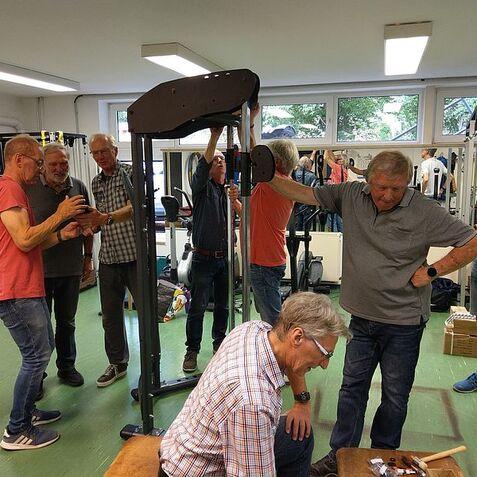 Gruppe älterer Männer montiert gemeinsam ein Fitnessgerät in einem gut beleuchteten Raum mit Sportgeräten.
