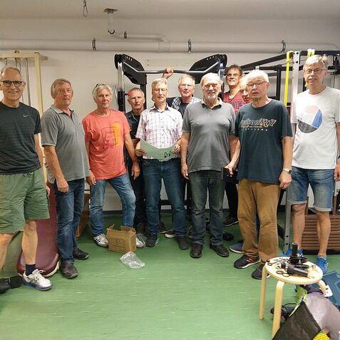 Gruppe von zehn Männern steht in einem Fitnessraum vor Trainingsgeräten, einige halten Unterlagen in den Händen.