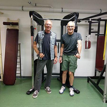 Zwei ältere Männer stehen in einem Fitnessraum vor Trainingsgeräten und halten jeweils einen Griff in der Hand.
