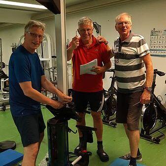 Drei Männer stehen in einem Fitnessraum neben einem Trainingsgerät und lächeln freudig in die Kamera.