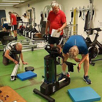 Drei Männer in einem Fitnessstudio testen Geräte, während sie auf blauen Matten und einem Trainingsgerät arbeiten.