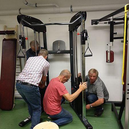 Drei Männer montieren gemeinsam ein Multifunktionsgerät in einem Fitnessraum. Ein Hocker steht im Vordergrund.