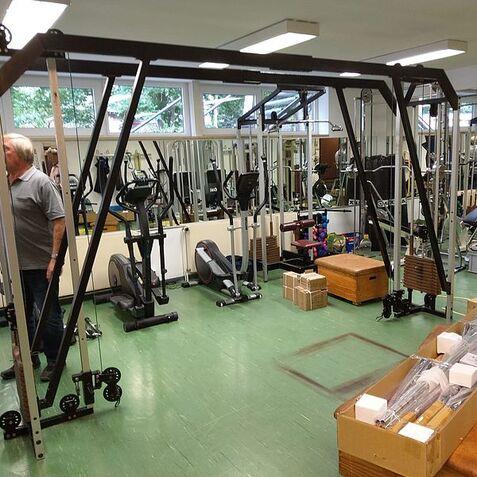 Fitnessraum mit einem Trainingsgerät, verschiedenen Trainingsgeräten und einem Mann im Hintergrund. Boxen sind auf dem Boden.