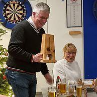 Mann hält einen Holzgegenstand bei einer Feier, umgeben von Menschen und Getränken an einem festlich dekorierten Tisch.