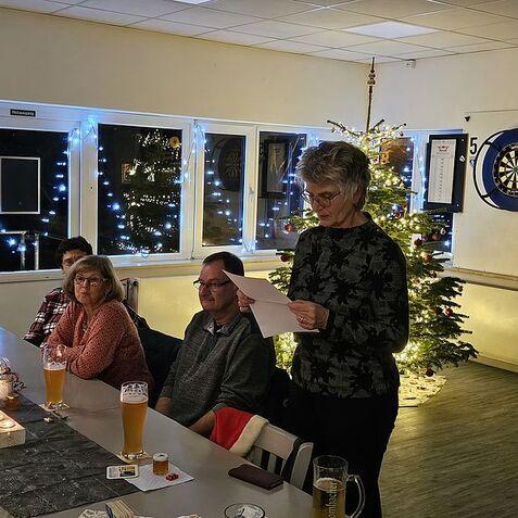 Frau liest an einem Tisch, umgeben von Gästen, mit Weihnachtsbaum und Lichtern im Hintergrund.