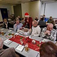 Weihnachtliche Feier mit gedecktem Tisch und Gästen, eine Person in rotem Hut serviert Speisen.