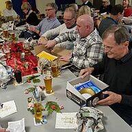 Personen sitzen um einen Tisch und öffnen Geschenke bei einer festlichen Veranstaltung, Getränke und Deko sind sichtbar.