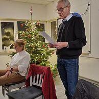 Mann liest vor einem Weihnachtsbaum, während eine Frau im Hintergrund auf einem Stuhl sitzt.