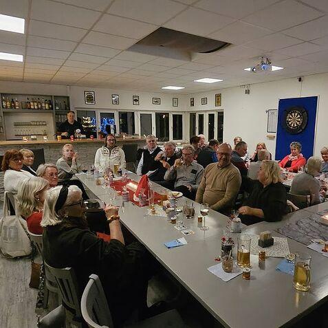 Gruppenszene in einem Raum mit Tisch, Getränken und Dartboards, Menschen sitzen und unterhalten sich entspannt.