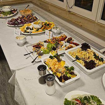Ein reichhaltiges Buffet mit Salaten, Fleisch und Beilagen auf einem weißen Tisch, beleuchtet von Fensterlicht.
