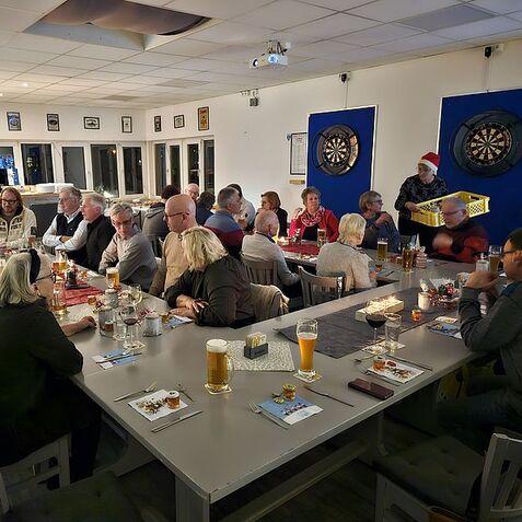 Gesellige Runde in einem Restaurant mit Menschen an langen Tischen, Getränken und Dartboards im Hintergrund.