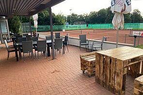 Terrasse eines Tennisclubs mit Holz- und Metallm&ouml;beln, Blick auf rote Tennispl&auml;tze im Hintergrund.