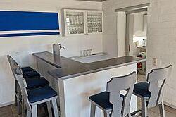 Moderne K&uuml;chenbar mit sechs blauen Hockern, &uuml;ber einem grauen Tresen, vor einer wei&szlig;en Wand mit blauer Akzentlinie.