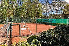 Blick auf einen Tennisplatz mit rotem Sand, umgeben von B&auml;umen und gr&uuml;nen Hecken, unter blauem Himmel.