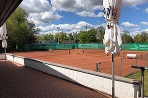 Tennispl&auml;tze mit rotem Sand und Sonnenschirmen unter wolkigem Himmel in einem gr&uuml;nen Umfeld.