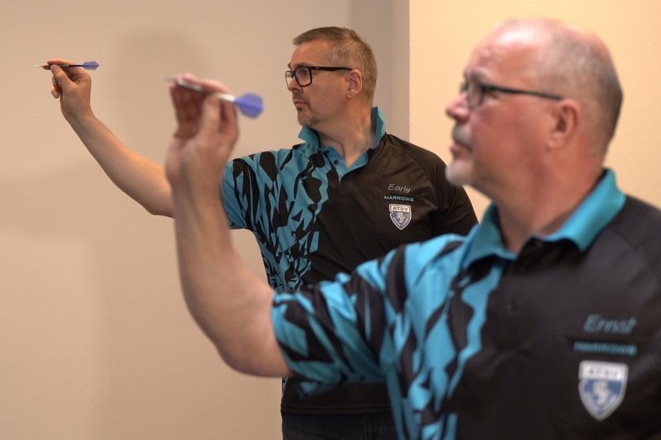 Zwei M&auml;nner in Dart-Shirts konzentrieren sich auf das Werfen von Darts in einem hellen Raum.